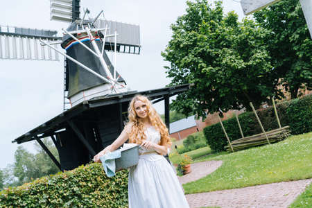 Woman dressed on traditional Dutch dress wooden shoes yellow clogs hanging Clothes on clothesline outside. laundry drying outdoor Netherlands windmill background on sun windy day. Retro vintage styleの写真素材