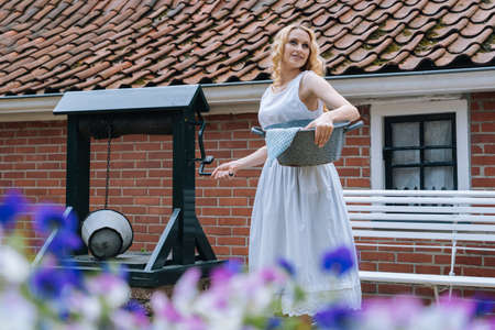 Woman in white dress holding basin near ancient artesian draw-well in village. Colorful violet flowers on foreground. Girl gets water from rural well in summer sunny day for washing.の写真素材
