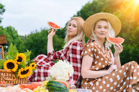 Friends is making picnic outdoor. Laughing girls sitting on white knit picnic blanket drinking wine. Happy young women talking, smiling outdoors on Summer picnic on sunny day at park.の写真素材