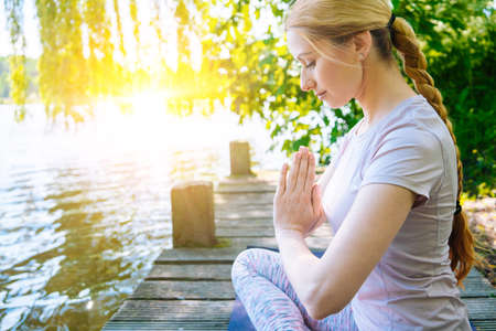 young woman doing yoga asana in park. girl stretching exercise in yoga position. happy and healthy woman sitting in lotus position and practicing yoga. meditation and sport on sunset outdoors.の写真素材