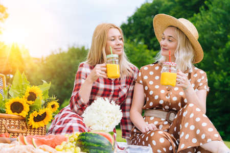 Friends is making picnic outdoor. Laughing girls sitting on white knit picnic blanket drinking wine. Happy young women talking, smiling outdoors on Summer picnic on sunny day at park.の写真素材