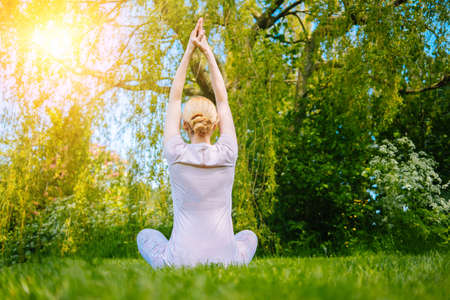 young woman doing yoga asana in park. girl stretching exercise in yoga position. happy and healthy woman sitting in lotus position and practicing yoga. meditation and sport on sunset outdoors.の写真素材