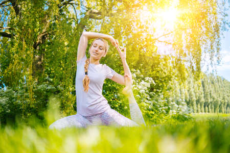 young woman doing yoga asana in park. girl stretching exercise in yoga position. happy and healthy woman sitting in lotus position and practicing yoga. meditation and sport on sunset outdoors.の写真素材