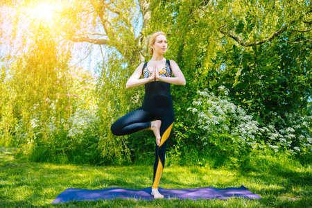 young woman doing yoga asana in park. girl stretching exercise in yoga position. happy and healthy woman sitting in lotus position and practicing yoga. meditation and sport on sunset outdoors.の写真素材