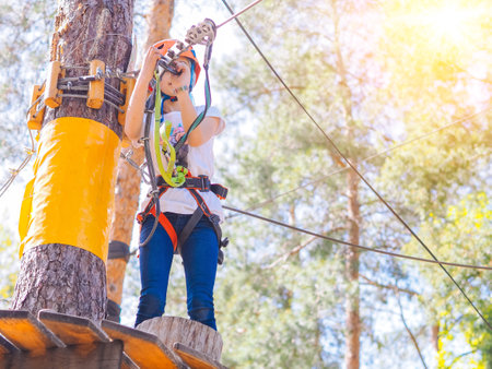 Kid in orange helmet climbing in trees on forest adventure park. Girl walk on rope cables and high suspension bridge in adventure summer city park. Extreme sport equipment helmet and carabinerの写真素材