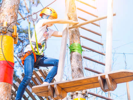 Kid in orange helmet climbing in trees on forest adventure park. Girl walk on rope cables and high suspension bridge in adventure summer city park. Extreme sport equipment helmet and carabinerの写真素材