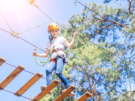 Kid in orange helmet climbing in trees on forest adventure park. Girl walk on rope cables and high suspension bridge in adventure summer city park. Extreme sport equipment helmet and carabinerの写真素材