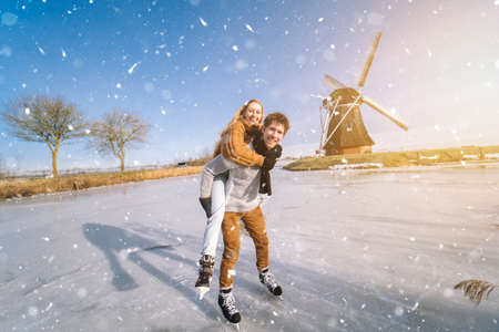 Loving couple having fun on ice in typical dutch landscape with windmill. Woman and man ice skating outdoors in sunny snowy day. Romantic Active date on frozen canal in winter Christmas Eveの写真素材