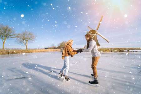 Loving couple having fun on ice in typical dutch landscape with windmill. Woman and man ice skating outdoors in sunny snowy day. Romantic Active date on frozen canal in winter Christmas Eveの写真素材