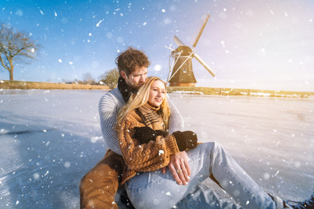 Loving couple having fun on ice in typical dutch landscape with windmill. Woman and man ice skating outdoors in sunny snowy day. Romantic Active date on frozen canal in winter Christmas Eveの写真素材