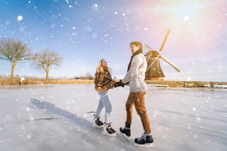 Loving couple having fun on ice in typical dutch landscape with windmill. Woman and man ice skating outdoors in sunny snowy day. Romantic Active date on frozen canal in winter Christmas Eveの写真素材