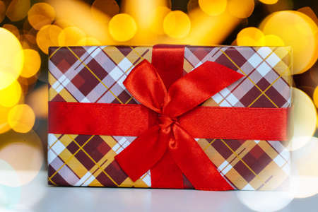 Christmas postcard with decorative gift box with red ribbon on light bokeh from Xmas tree. New Year and Christmas design. Holiday greeting card.の写真素材