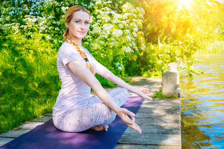 young woman doing yoga asana in park. girl stretching exercise in yoga position. happy and healthy woman sitting in lotus position and practicing yoga. meditation and sport on sunset outdoors.の写真素材