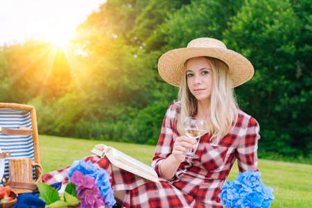 Girl in red checkered dress and hat sitting on white knit picnic blanket reading book and drinking wine. Summer picnic on sunny day with bread, fruit, bouquet hydrangea flowers. Selective focus.の写真素材