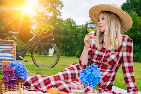 Girl in red checkered dress and hat sitting on white knit picnic blanket reading book and drinking wine. Summer picnic on sunny day with bread, fruit, bouquet hydrangea flowers. Selective focus.の写真素材
