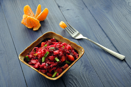 Bowl of russian salad vinaigrette of beetroot, cabbage, carrot, potatoe and green onion, with hulled mandarin on the fork on the blue wooden table, selective focusの写真素材