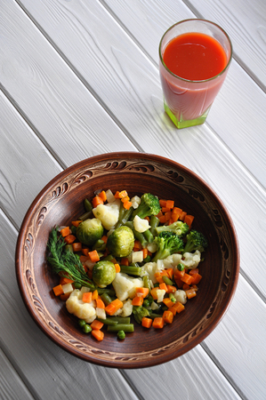 Vegetable ragout made of cauliflower, broccoli, carrot, Brussels sprouts, green pea and fresh dill served in clay dish with tomato juice on the white wooden table. Selective focus.の写真素材