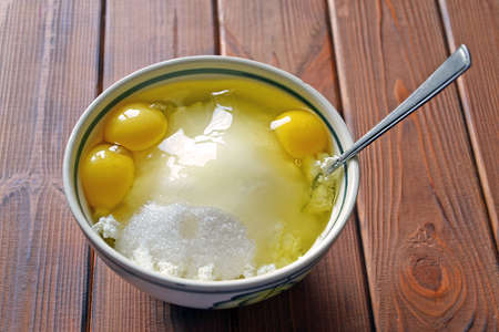 White ceramic bowl with three yolks, sugar and cottage cheese - preparing pastry at home. Selective focus on the wooden backgroundの写真素材