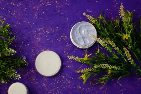 Face or body care cream in white container. Organic natural herbal cosmetics, moisturizer for skin. Flowers on purple background. Top view, copy space.の写真素材