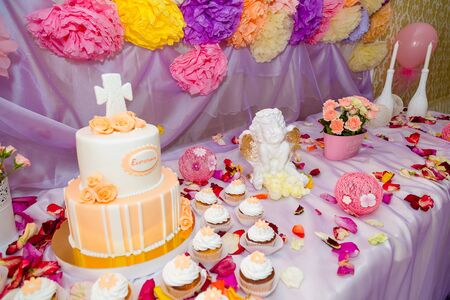 Baptism candy bar, angel statue, cupcakes, unfocused cake. Mastic cross on cake for christening child party. White candles and flower petals on the table.の写真素材