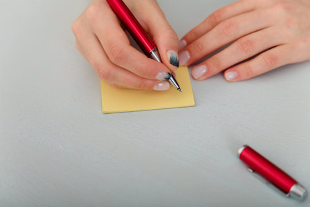 Female hands with fir-tree forest manicure holding red pen over yellow paper stickers. Christmas or...