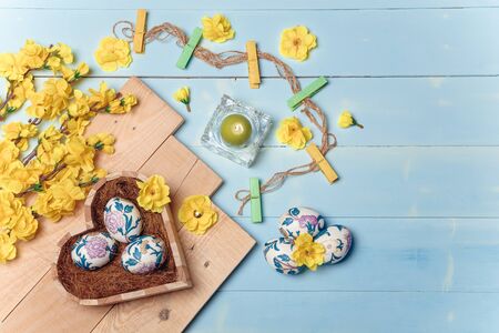 Heart shaped box with decorated Easter eggs, yellow flowers, burning candle and brown boards on wooden blue background. Spring and Easter concept. Top view, flatlay, copy space.の写真素材