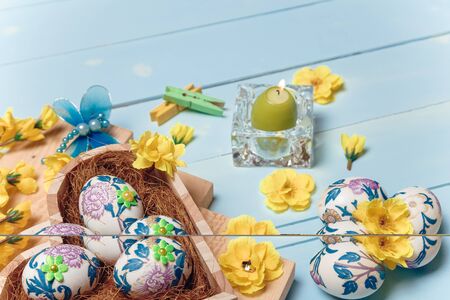 Heart shaped box with decorated Easter eggs, yellow flowers, burning candle and brown boards on wooden blue background. Spring and Easter concept. Close up, copy space.の写真素材