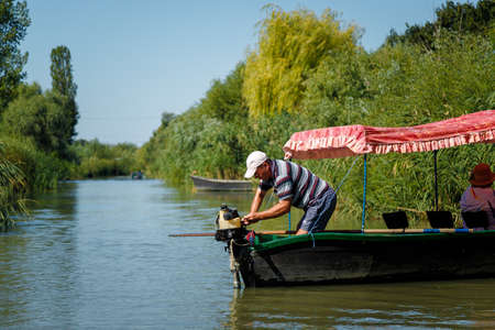 Vilkovo, Ukraine - August 2019. Man turning on a motor boat engine at river or lake among green trees. Contryside summer landscape, leisure fishing concept.のeditorial素材