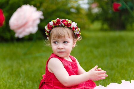 Little girl in red dress playing  on the green lawn with high artificial rose flowers on background. Springtime, blossom and childhood concept.の写真素材