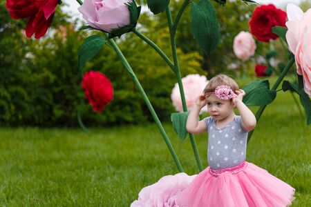 Little girl in pink fluffy tutu skirt playing  on the green lawn with high artificial rose flowers on background. Springtime, blossom and childhood concept.の写真素材