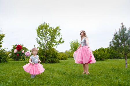 Little girl and mother in pink fluffy tutu skirt playing  on the green lawn with high artificial rose flowers on background. Springtime, blossom and childhood concept.の写真素材