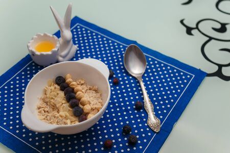 Oatmeal porridge in white bowl with blueberries, almond flakes, nuts. Pot of honey and spoon on blue napkin on kitchen table. Healthy breakfast and nutrition concept.の写真素材