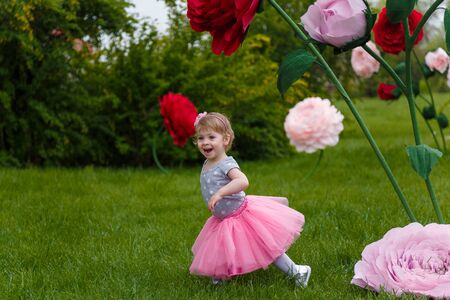 Little girl in pink fluffy tutu skirt playing  on the green lawn with high artificial rose flowers on background. Springtime, blossom and childhood concept.の写真素材