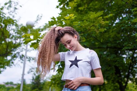 Teenage girl in white T-shirt with star design holding ponytail of long curly hair walking in green parkの写真素材