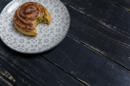 Bitten cheese and dill bread bun on plate standing on black wooden background. Tasty unhealthy food....