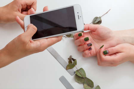 Nail artist taking photo with smartphone of her work, clients manicured hands. Green nail polish design, nail files and eucalyptus branch on white table. Spring manicure.の写真素材