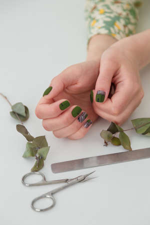 Female hands with green manicure and geometric nail design. Scissors, nail files and eucalyptus branches on white table. Spring nail art, beauty concept. Vertical shot, copy space.の写真素材
