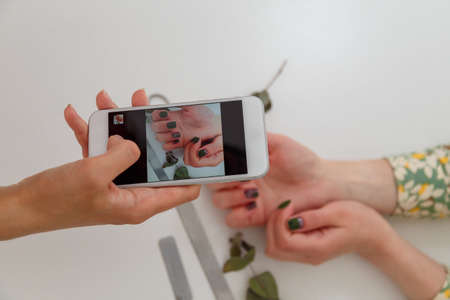 Nail artist taking photo with smartphone of her work, clients manicured hands. Green nail polish design, nail files and eucalyptus branch on white table. Spring manicure. Copy space.の写真素材