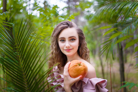 Teenager girl with curly long hair in pink costume among green trees and bushes holding exotic fruit grapefruitの写真素材