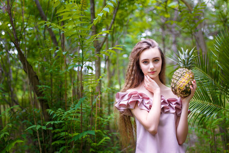 Teenager girl with curly long hair in pink costume among green trees and bushes holding exotic fruit pineappleの写真素材