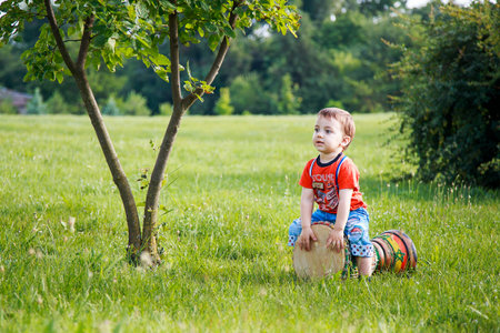 Little boy in green park sitting on bongo or tomtom drum and playing musical instrument. Carefree childhood and outdoor summer leisure concept.の写真素材