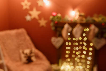 Unfocused shot. Blurry Christmas background with arm-chair and fireplace decorated with hanging socks, fir branches, red baubles and golden garland lights.の写真素材