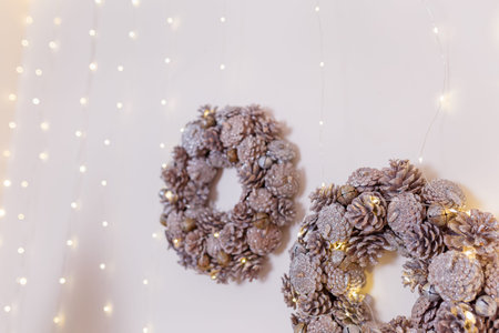Two Christmas wreath made from pine cones with shiny garland lights hanging on white wall, copy...