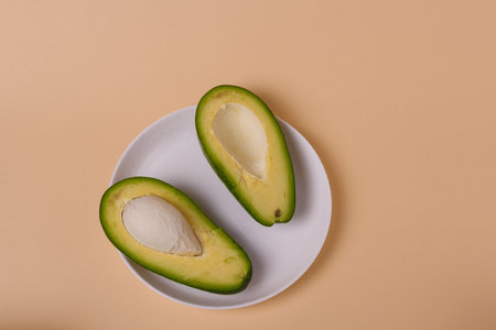 Two halves of green fresh avocado with seed on white plate on beige background. Minimal styled...