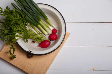 Green onions, parsley, radish in white metallic bowl on kitchen cutting board on wooden table. Fresh vegetables and greenery, organic vegetarian food concept. Top view, copy space.の写真素材