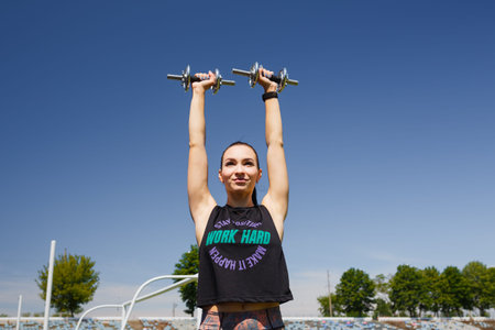 Smiling girl standing on stadium green lawn with dumbbells in hands. Sport, physical training,...