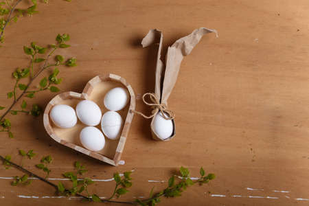 Heart shaped box with boiled eggs and Easter bunny made from egg and paper ears with green branch on wooden brown background. Easter and spring concept. Top view, copy space.の写真素材