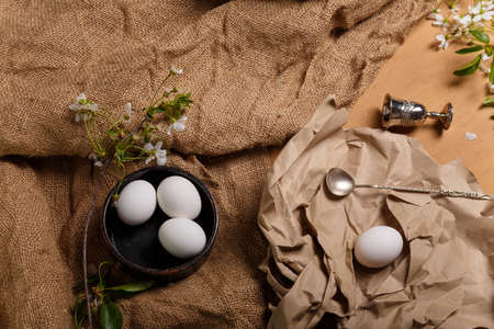 Easter eggs in black bowl, silver spoon, egg cup, cherry blossom on paper and sackcloth on wooden brown background. Easter and spring concept, top view.の写真素材