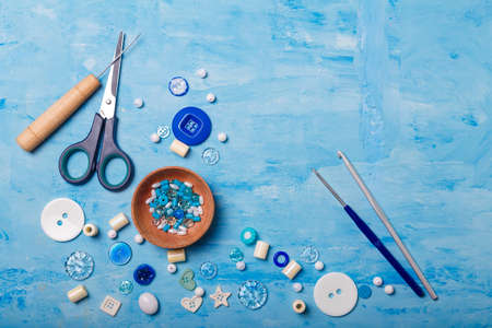 Close up of wooden bowl of beads and buttons, awl, scissors and crochets for sewing and embroidery. Set of materials for handcraft, making of bijouterie and accessories. Blue background, copy space.の写真素材