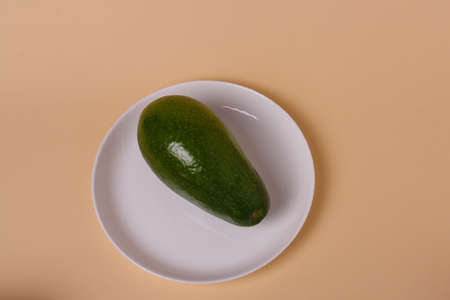 One whole green fresh avocado on white plate on beige background. Minimal styled composition.の写真素材
